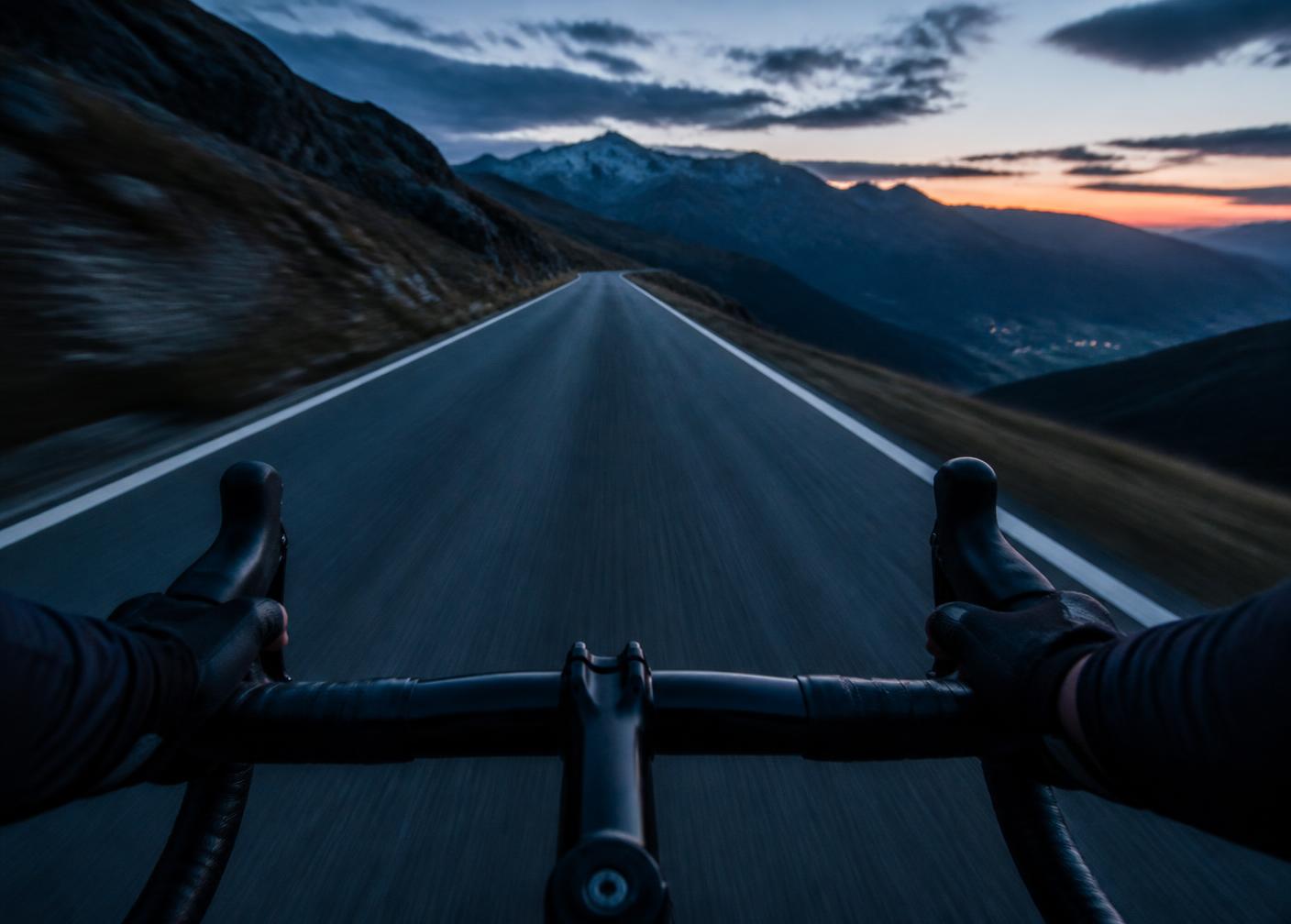 Blick aus Fahrerperspektive über den Rennradlenker auf eine Bergstraße in der Dämmerung
